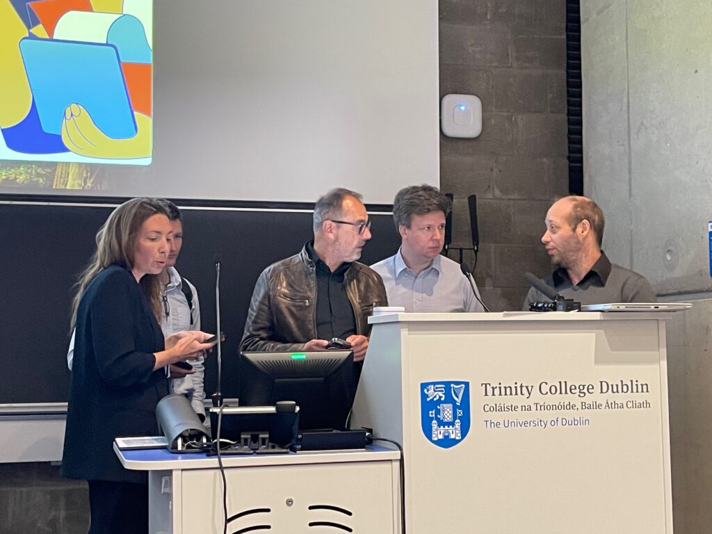 5 personnes au pupitre dans un amphithéatre, la figure de l'une d'entre-elles est cachée. Devant le pupitre la mention Trinity College Dublin est inscrite, accompagnée d'un écusson. 